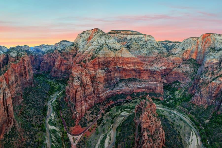 Those who climb the dangerous trail Angels Landing are rewarded at the top with a fantastic view over Zion National Park and the reddish rocks. The picture shows the river below, winding through the canyon, the red rocks, and a pink sunset in the sky.