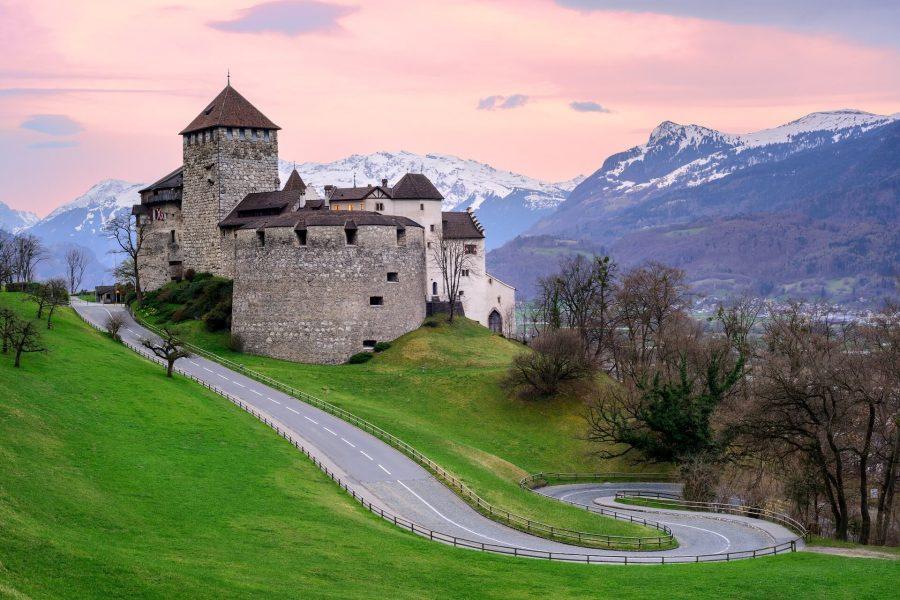 Vaduz Castle in Liechtenstein, a fortress with thick stone towers, sits atop a green hill with mountains in the background.