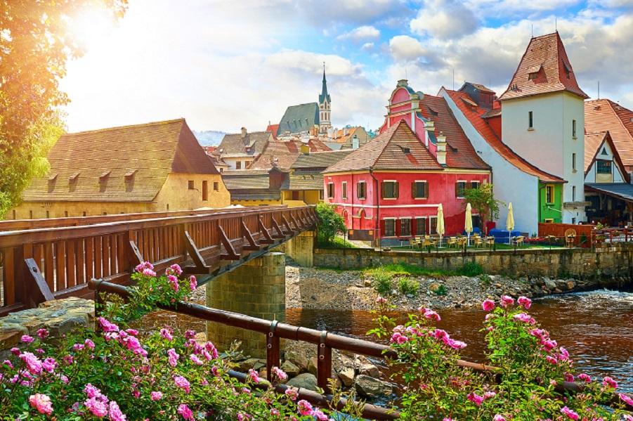 Czech Krumlov, (Cesky Krumlov), Czech Republic. Wooden bridge over the Vltava River. Vintage picturesque old town with colorful houses and a church chapel. Rose flowers on a bench. Sunny summer day.