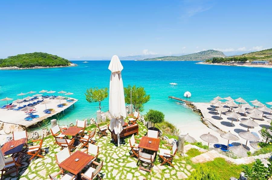 The beach of Saranda in Ksamil, Albania, is one of the most beautiful spots in the country. In the foreground are wooden tables and a sun umbrella. Sun loungers line the beach, and the water in the background is clear and a vibrant turquoise.