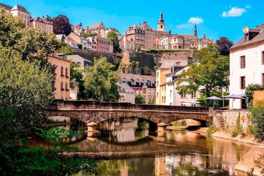 The idyllic valley along the Alzette River in Luxembourg features many charming houses, and a low stone bridge with several arches spans the river.