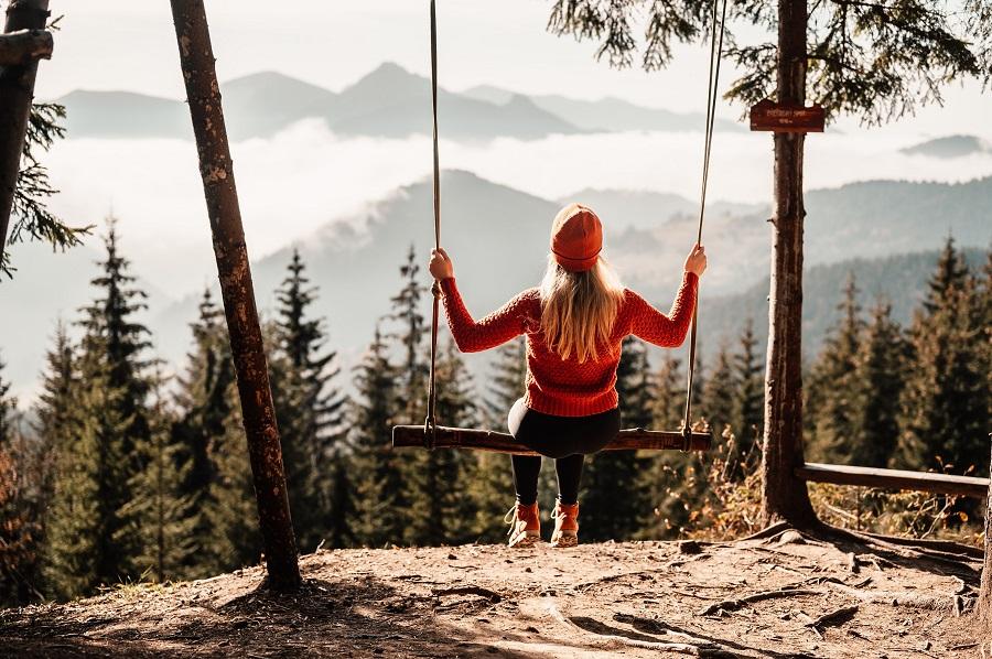 A woman sits on a swing on a mountain overlooking a dense coniferous forest and misty mountains.
