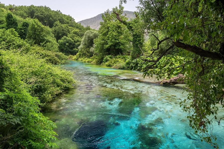 Crystal clear blue-green water of Syri i Kalter in Albania, surrounded by lush greenery. In the background, wooded hills rise. The river flows from the bottom left to the top right.