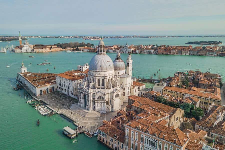 Aerial view of Santa Maria della Salute. It is surrounded by water on the left and right. It stands out particularly because two large white domes rise into the sky.