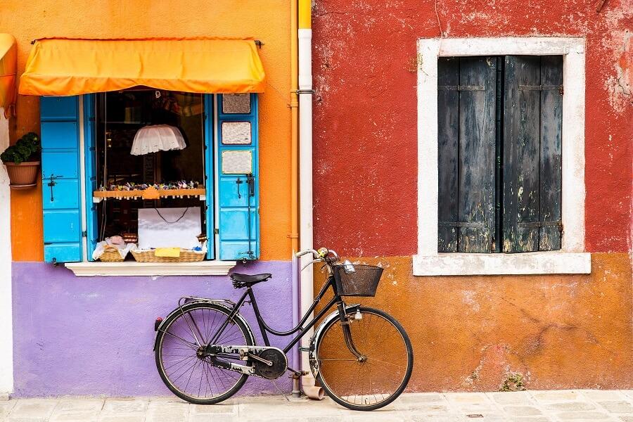 A colorful bicycle leans against a vibrant house wall in Burano. On the left, a light blue window with an orange awning; on the right, a dark window in front of a reddish-brown wall. Cobblestones form the ground.