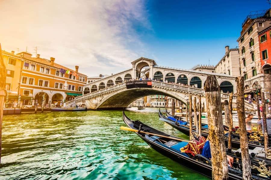 The focus of the picture is the Rialto Bridge in Venice, which spans the Grand Canal. It reaches its highest point above the canal in the middle. In the foreground, you can see some gondolas floating in the water on the right.