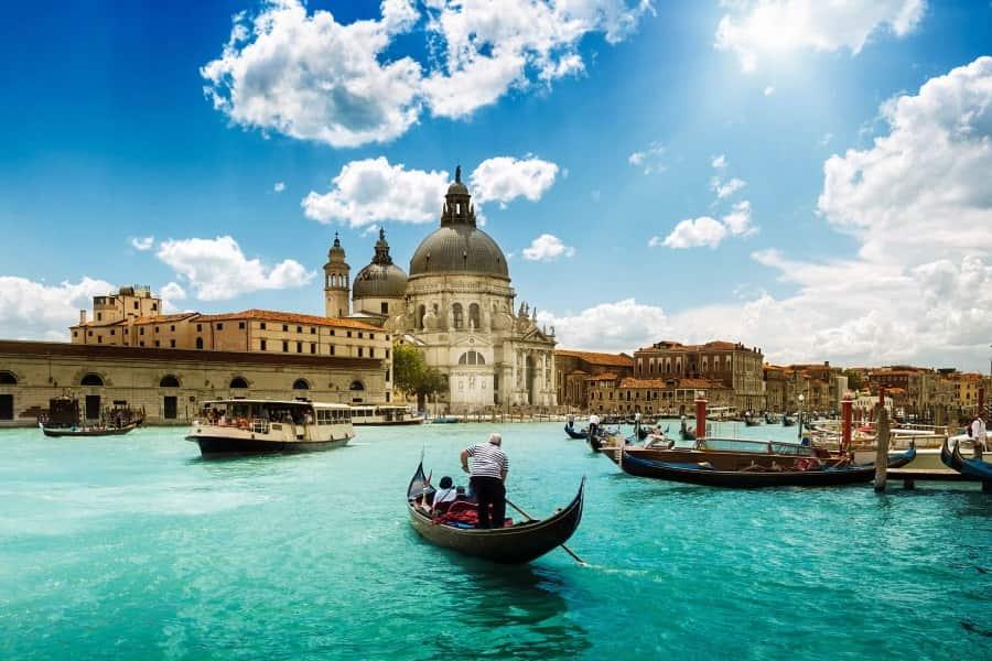 Gondolas on radiant blue water in Venice, with a large church in the background under a clear blue sky. Historic buildings line the shore, and the scene looks picturesque and lively.
