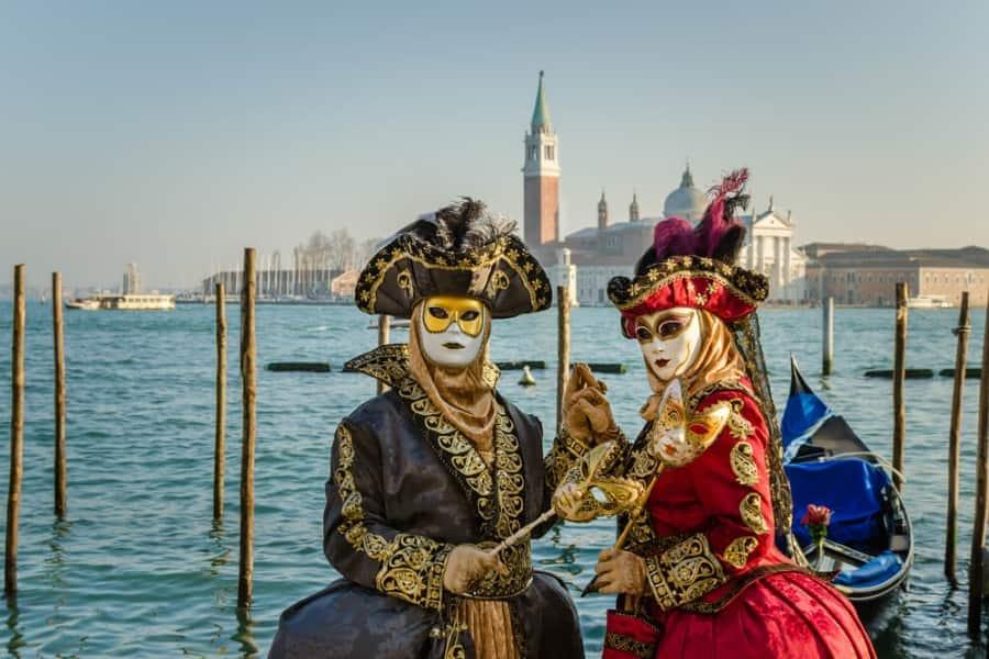 Two costumed individuals in Venice stand in front of the water holding hands. The two people wear identical outfits: a lavish dress with golden embellishments, a large feathered hat, and a golden mask over their eyes. The dress of the left person is black; that of the right is red.