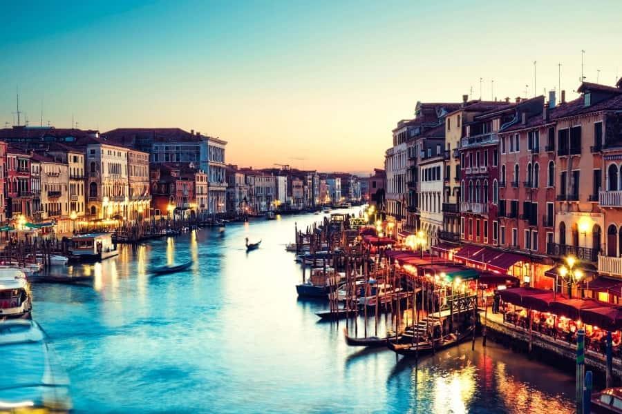 The image shows the Grand Canal in Venice at dusk. Along the edges, gondolas are moored, behind them the lined-up restaurants with their lights already on.