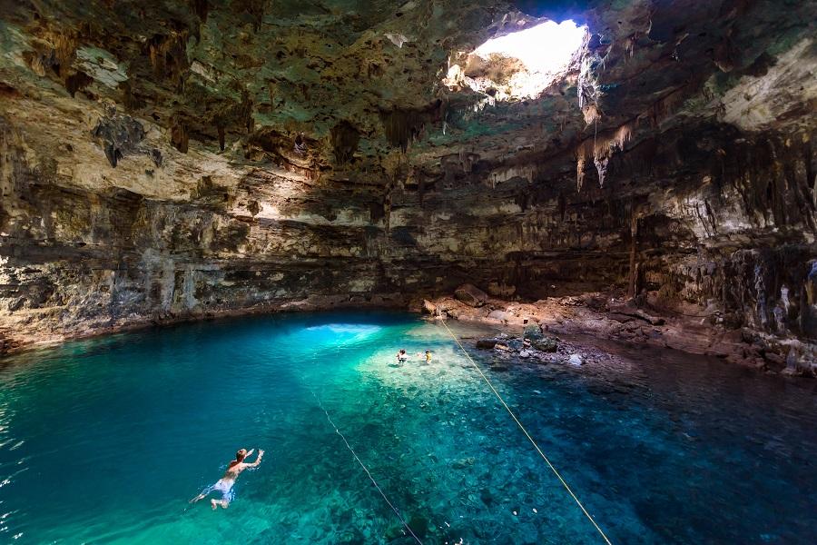 People swimming in clear, turquoise water in a cave. Sunlight falls through a hole in the ceiling and illuminates the area. Stalactites hang from the rocky ceiling.