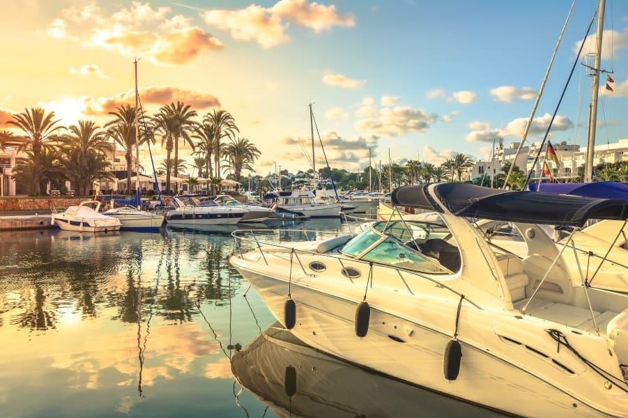 The harbor of Cala d'Or at sunset. In the foreground, on the right side, you can see a speedboat. In the background, more white boats can be seen lying before a row of palm trees in the water. Above the palms, the sky is turning yellow-red.