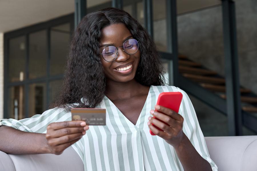 A young woman sits on a sofa, smiling, holding a smartphone in her right hand. In her left hand, a no-foreign-transaction-fee credit card. Behind her, modern architecture with glass and stairs.
