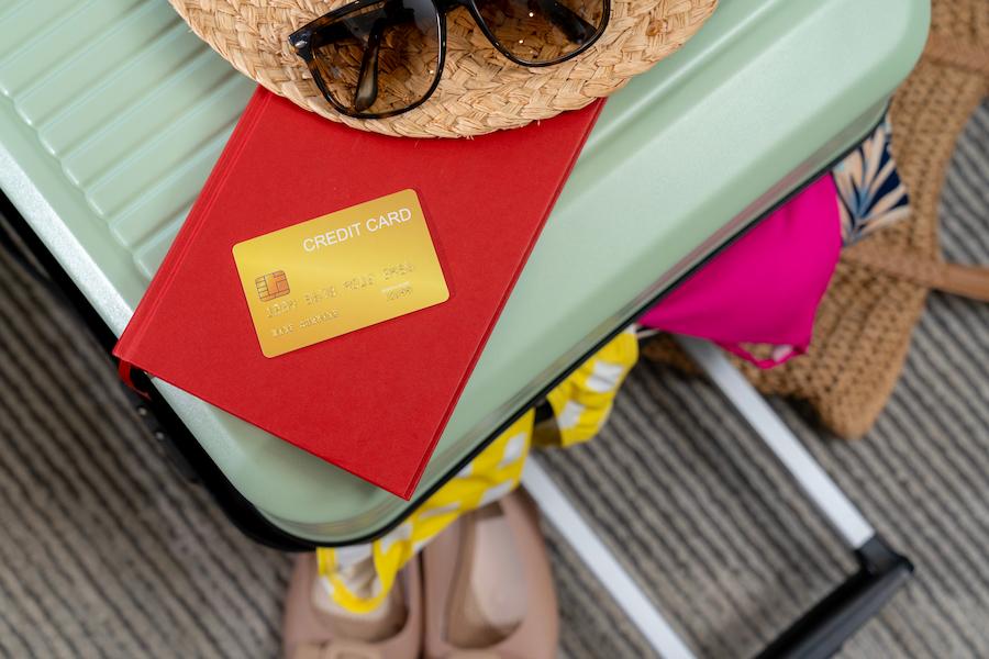 On a green suitcase lie a red book, a no-foreign-transaction-fee credit card, and a straw hat with sunglasses. Next to it are colorful clothes.