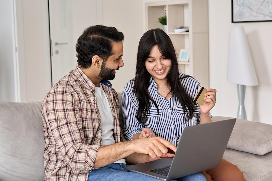 A smiling couple sits on a sofa, the man points at a laptop while the woman holds a no-foreign-transaction-fee credit card. In the background, there is a shelf and a floor lamp.