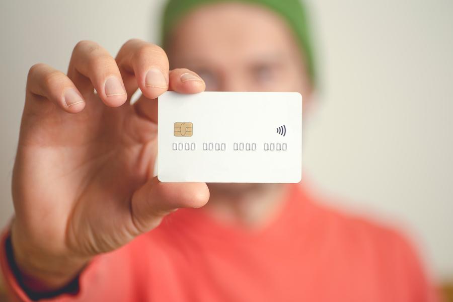 Close-up of a hand holding a white credit card. There is a gold chip at the top left and a contactless payment symbol on the right. The card could be a credit card without foreign transaction fees.