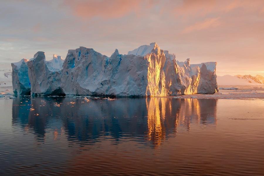 Iceberg at sunset reflecting golden light, in front of calm water in Antarctica. Pink-orange clouds in the sky and sun reflections in the water create a peaceful atmosphere.
