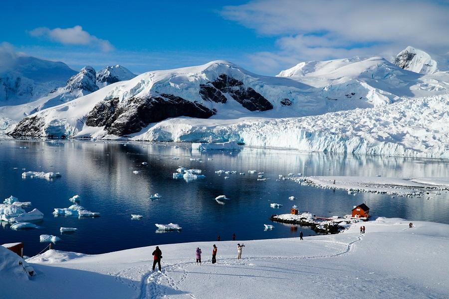 Snow-covered landscape with people in the foreground. Icebergs in the sea on the left, a small cabin by a dock on the right. Majestic, snow-covered mountains under a blue sky in the background.