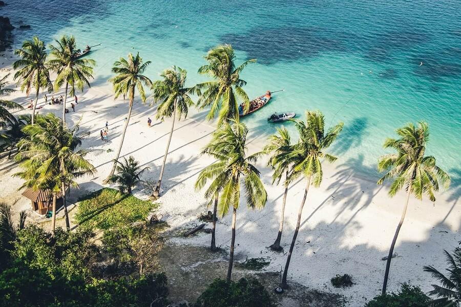Sunny beach with turquoise water lined by tall palm trees casting long shadows on the white sand. Boats are in the water near the shore, people walk along the palms.