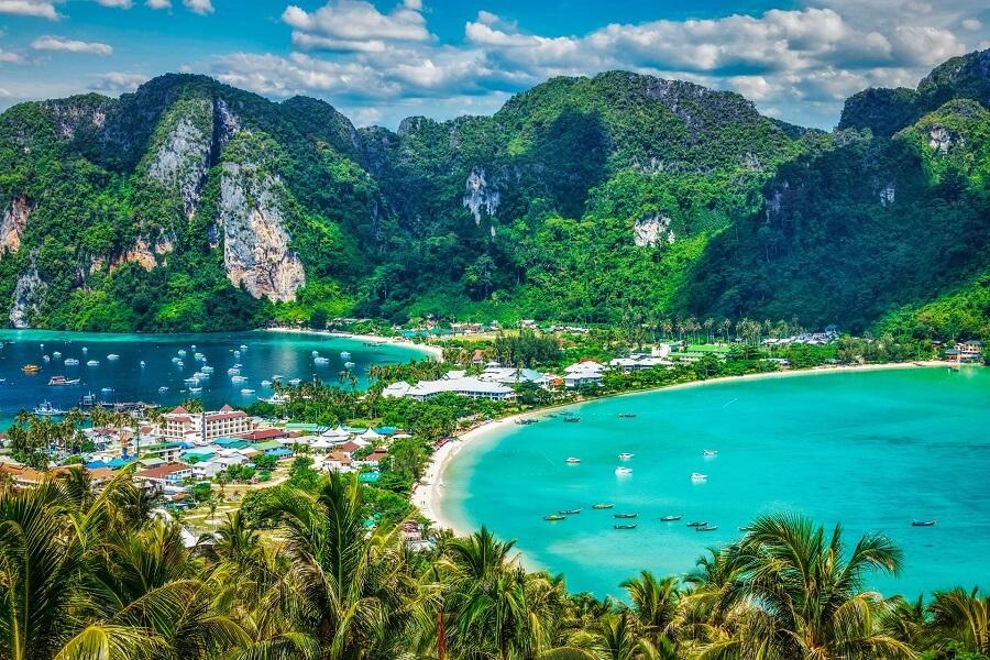 Lush green mountains frame two bays with turquoise water and white sandy beaches lined with trees in the foreground and numerous boats on the water. A lively coastal settlement lies in between.