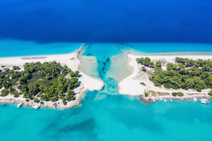 Bright blue sea separates two green peninsulas with light sandy beach. In the foreground, turquoise water meets the shore, while dense trees line the scenery.