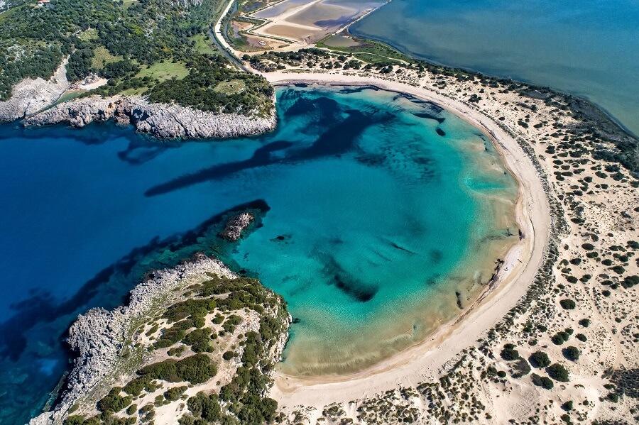 Aerial view of a crescent bay with turquoise water and sandy beach, surrounded by green vegetation. Rocky cliffs to the left, in the background the blue sea.