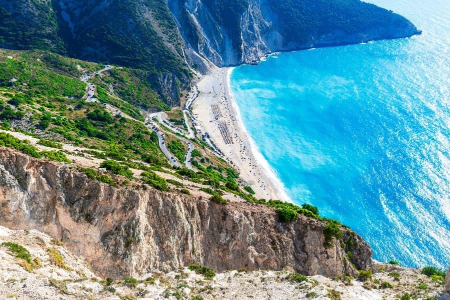 Steep cliffs at Myrtos Beach on Kefalonia, Greece, lead to a white sandy beach washed by turquoise water. Lush greenery covers the slopes in the background.