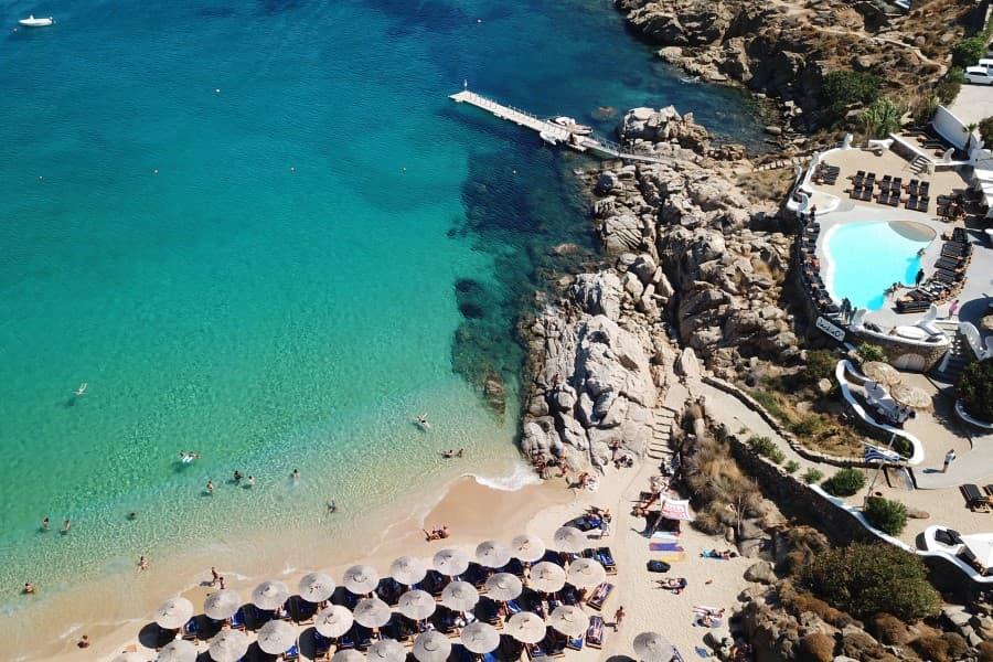 White sandy beach with sun umbrellas in front, turquoise water on the left. To the right, rocky coast with a round blue pool facility. Scattered bathers in the sea and on land.