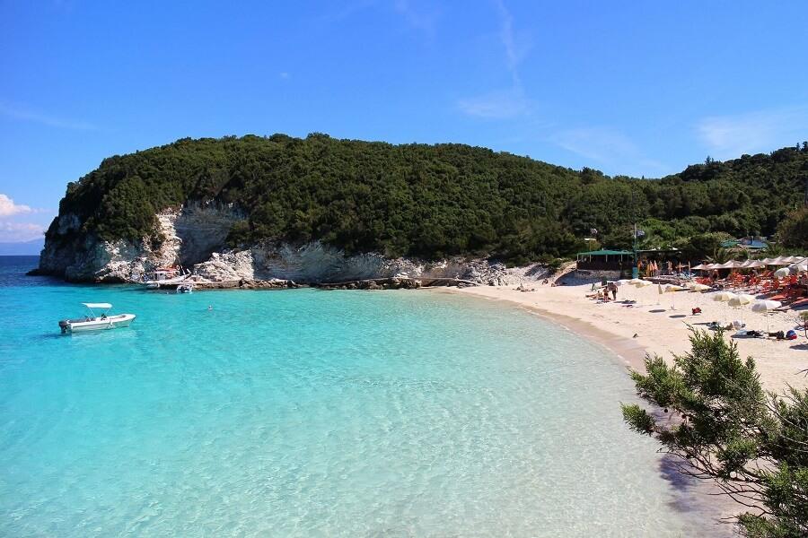 Clear turquoise sea, sandy beach with sunbeds and umbrellas. Right, tavernas under trees, left, a boat floats. In the background wooded cliffs under a blue sky.