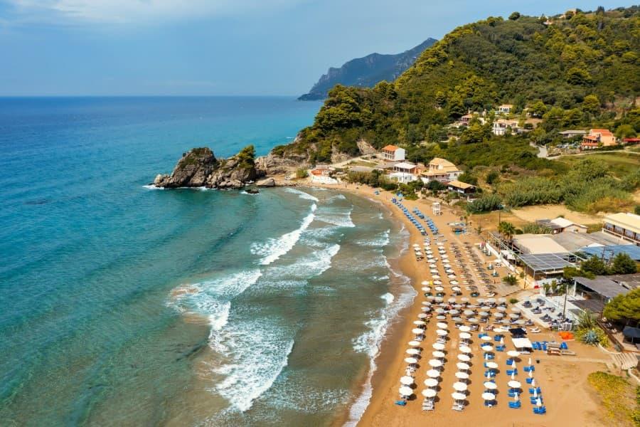 Fine sandy beach with rows of white umbrellas and sunbeds, turquoise sea with gentle waves on the left, green hills and holiday homes on the right, blue sky in the background.