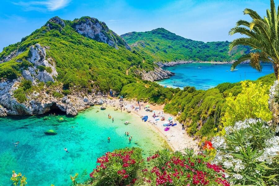 Aerial view of Porto Timoni in Corfu: Two turquoise bays, between them a green peninsula. Beaches visited by people, surrounded by lush vegetation and palm trees in the foreground.