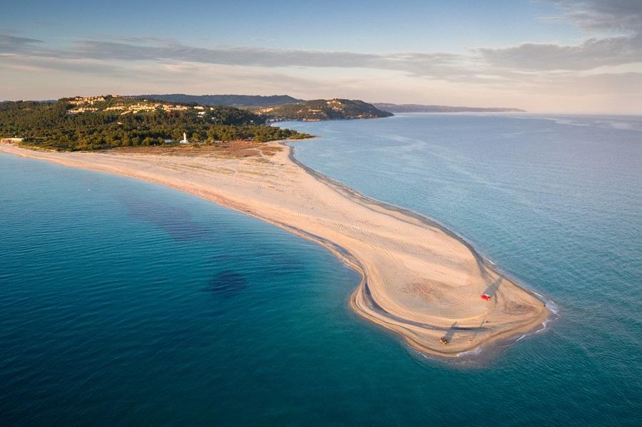 A narrow sandy strip extends into the blue sea, surrounded by water. On the left, dense forest, in the background hilly landscape. The right side of the beach is slightly curved, with a small dune visible.