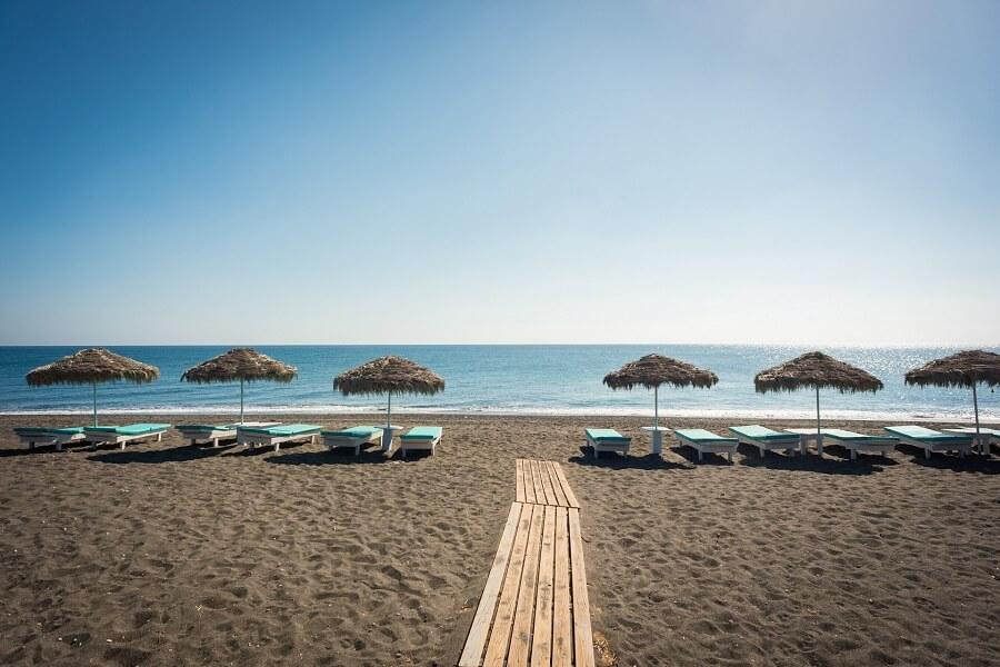 Beach with black sand, several blue sunbeds and thatched roofs. A wooden path leads through the sand. Clear view of the calm blue sea and a cloudless sky.