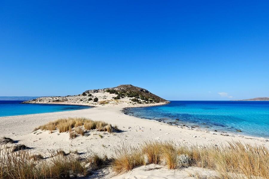 Clear turquoise water surrounds the sandy Simos Beach on the island of Elafonisos in Greece. In the foreground small tufts of grass grow, in the background a green island rises.