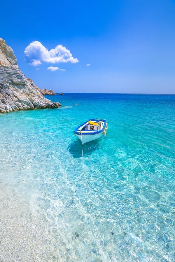 A small boat floats on crystal clear turquoise water near a rocky shore on the left. The sky is a bright blue with a few clouds. A rope connects the boat to the shore.