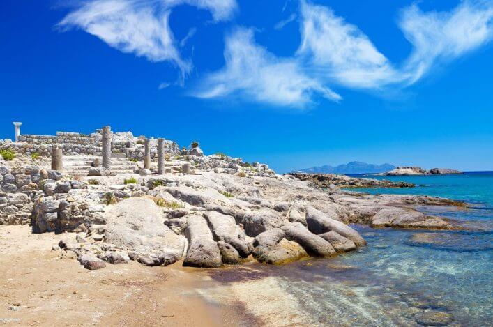 Ruins of the Agios Stefanos Basilica on Kos, Greece. In the foreground a sandy beach, behind ancient columns and ruins, surrounded by clear blue water. Blue sky with white clouds.