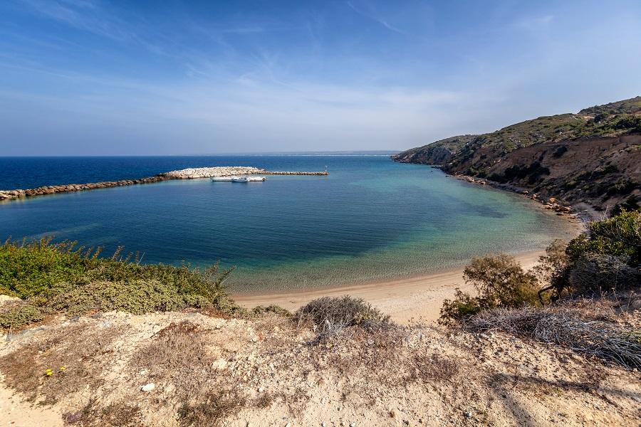 Sandy beach in front of clear blue sea, surrounded by a long jetty on the left and wooded hills on the right. Under a blue sky, the landscape looks calm and inviting.