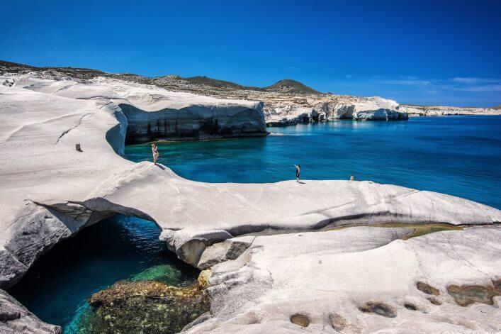 White, smoothly polished rocks extend alongside the turquoise sea, interrupted by small coves. People stand on the rocks while the azure sky crowns the scene.