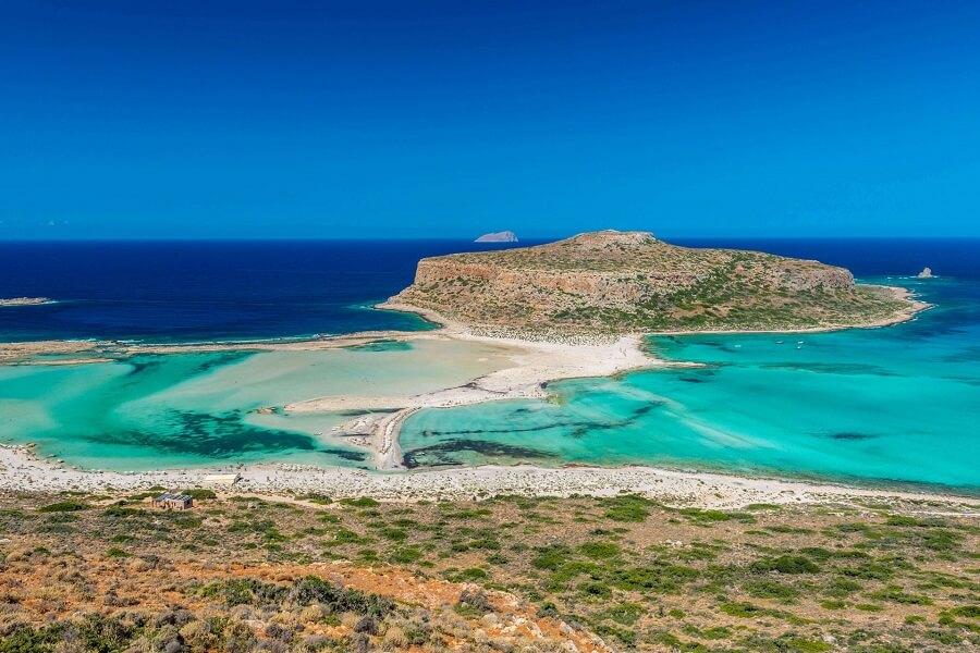Clear turquoise water, surrounded by a sandy lagoon in the middle. In the background, a green island under a blue sky. The foreground features dry, grassy coastal rocks.