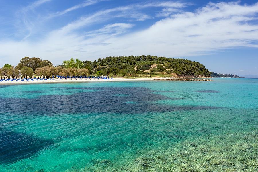 Bright blue sea separates two green peninsulas with light sandy beach. In the foreground, turquoise water meets the shore, while dense trees line the scenery.