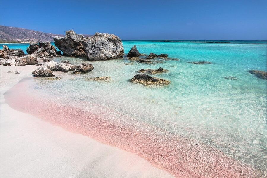 Gentle waves meet a pink-tinged sandy beach with clear turquoise water. In the background, rocky cliffs rise, creating an idyllic coastal section of Crete.