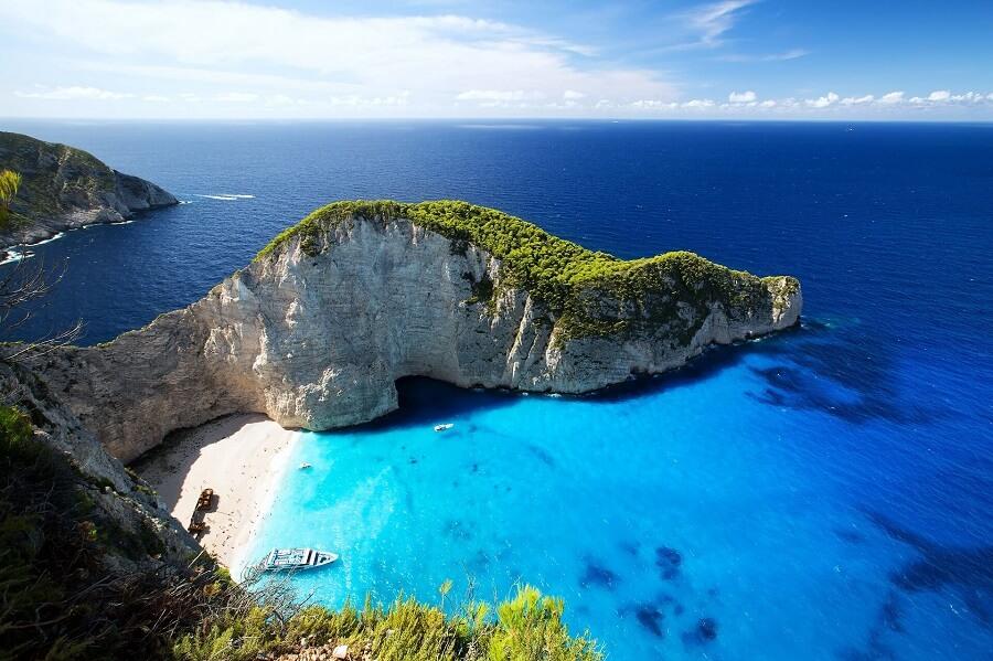 An idyllic bay with turquoise water and white sand in a rocky cove. On the left, a stranded ship, on the right, green cliffs, topped by a clear blue sky.