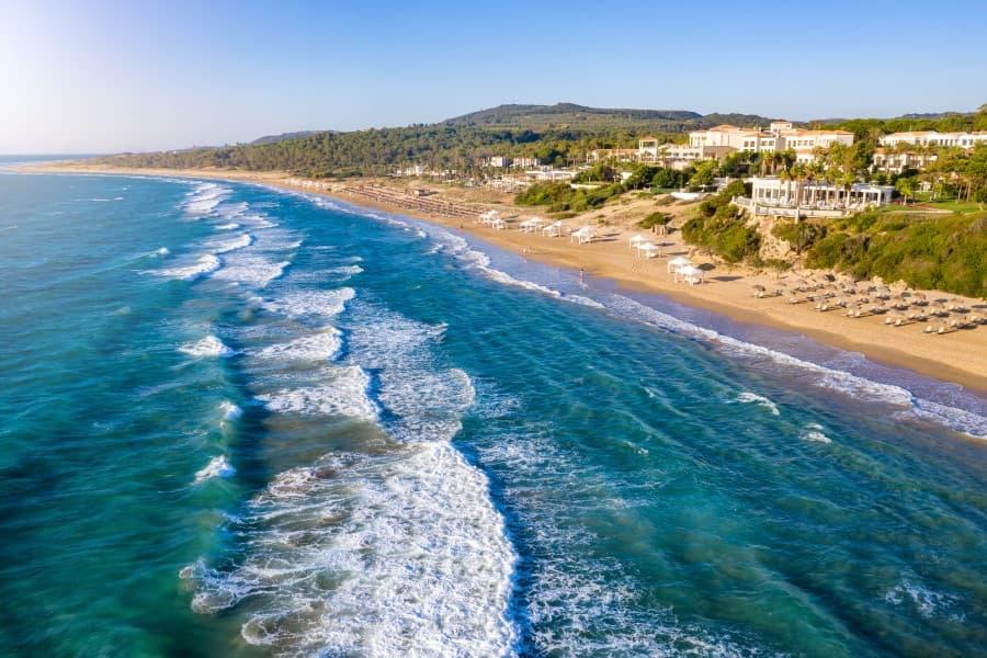 Waves lap the wide sandy beach of Kyllini Beach, Peloponnesus, lined with sun umbrellas and loungers on the right, while green hills and buildings are visible in the background.