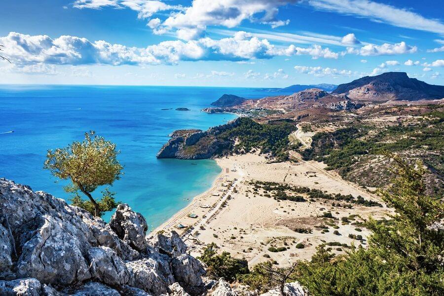 View from Tsambika Monastery in Rhodes: Turquoise sea on the left, golden sandy beach in the center, surrounded by green hills on the right. Scattered clouds and rocks in the foreground.