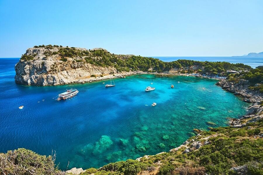 Turquoise sea in a cove, surrounded by green hills and cliffs. Several boats float on the water. In the background, the landscape fades into the blue sky.