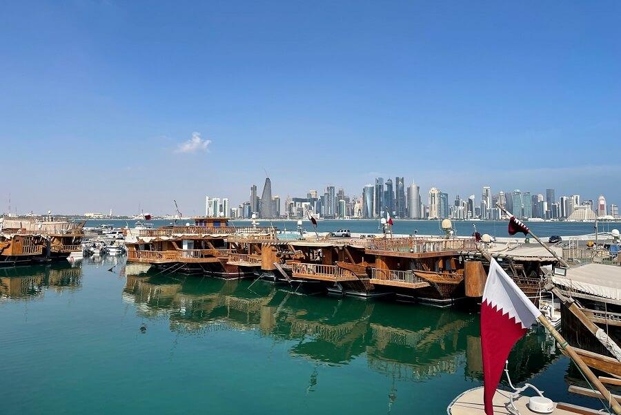 Traditional wooden boats float in the clear waters of Qatar's Dhow Harbour. The impressive skyline of Doha rises in the background. A Qatari flag flutters in the foreground.