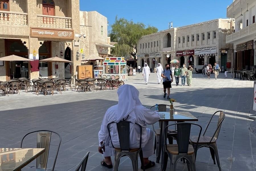 In the Souq Waqif in Qatar, a person in white attire sits at a café table in the foreground, surrounded by empty chairs. In the background, traditional buildings and strolling people can be seen.