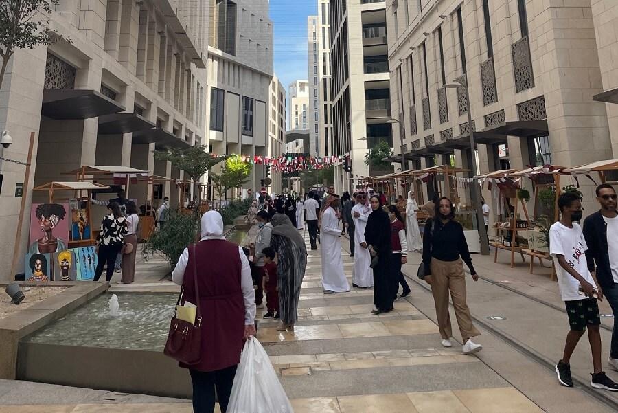 People stroll down a busy street in Msheireb, Qatar, lined with tall, modern buildings. In the foreground are stalls selling artwork and a small water oasis. Ideal for Qatar travel tips.