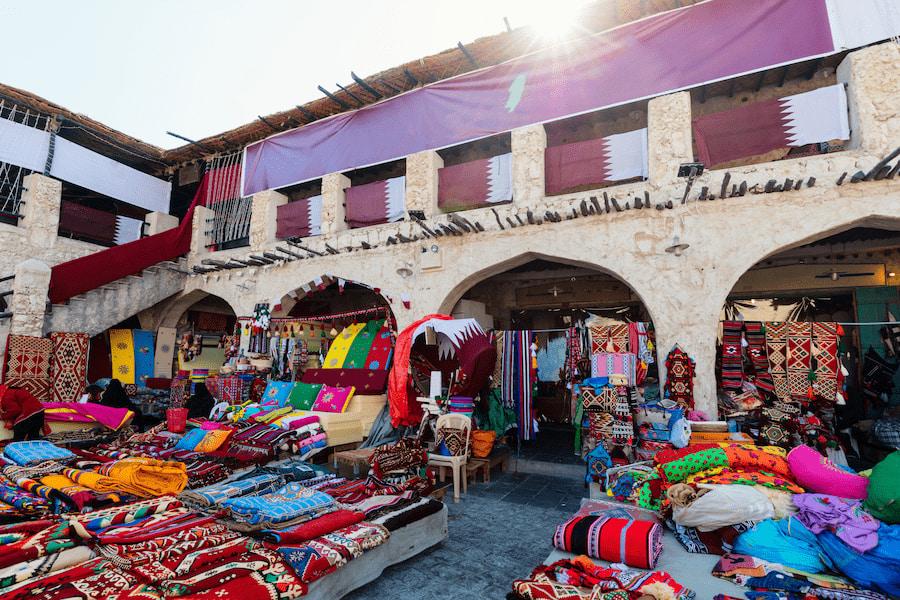 Colorful rugs and cushions are stacked on a market stall in Doha, Qatar. Traditional textiles and decorations adorn the area, surrounded by historical stonework and arches.