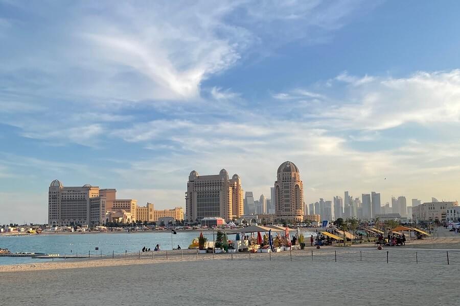 Beach with a view of Qatar's impressive skyline. Large, distinctive buildings on the left and center. In the foreground, colorful umbrellas and a waterway. An ideal tip for a visit to Qatar.