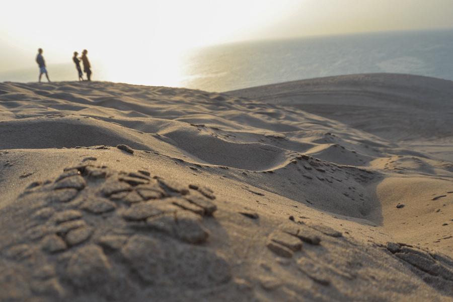 Desert landscape in Qatar: Three people hike in the background across gentle dunes near Chaur al-Udaid. The sun is low over the sea, casting soft light on the sand.